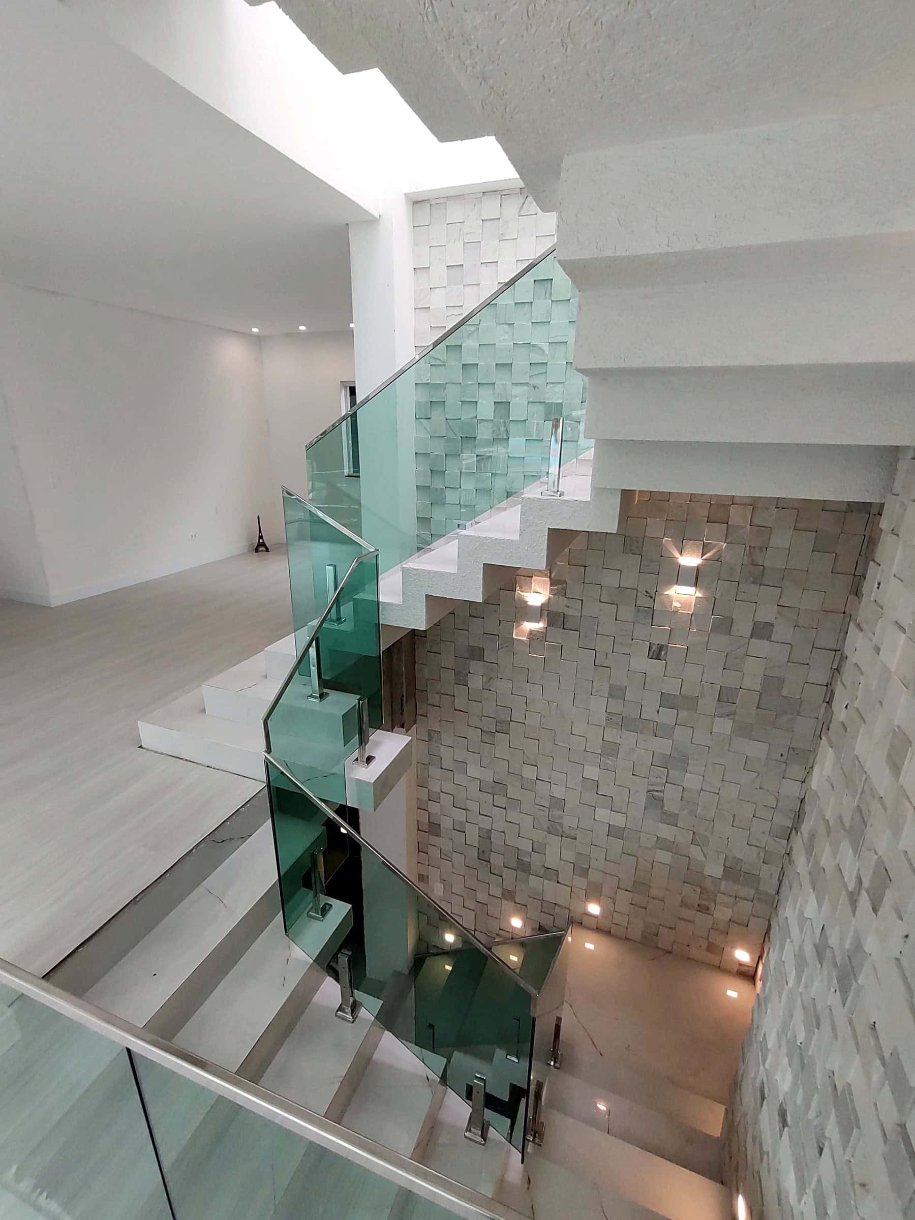 Escada de três pavimentos vista do alto, com guarda-corpo de vidro verde e colunas em inox ao lado de parede de pedra