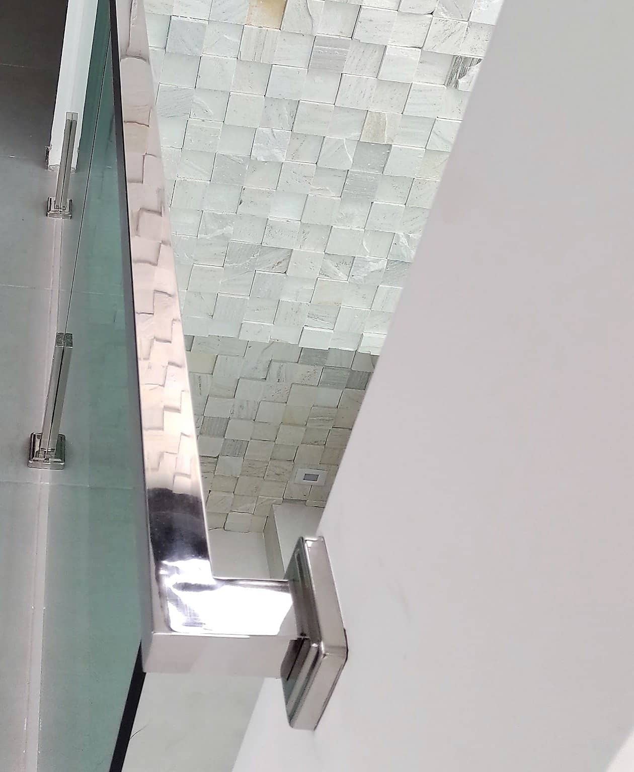Detalhe de corrimão em inox polido sobre vidro verde, refletindo a parede de pedra, com o suporte fixado na parede
