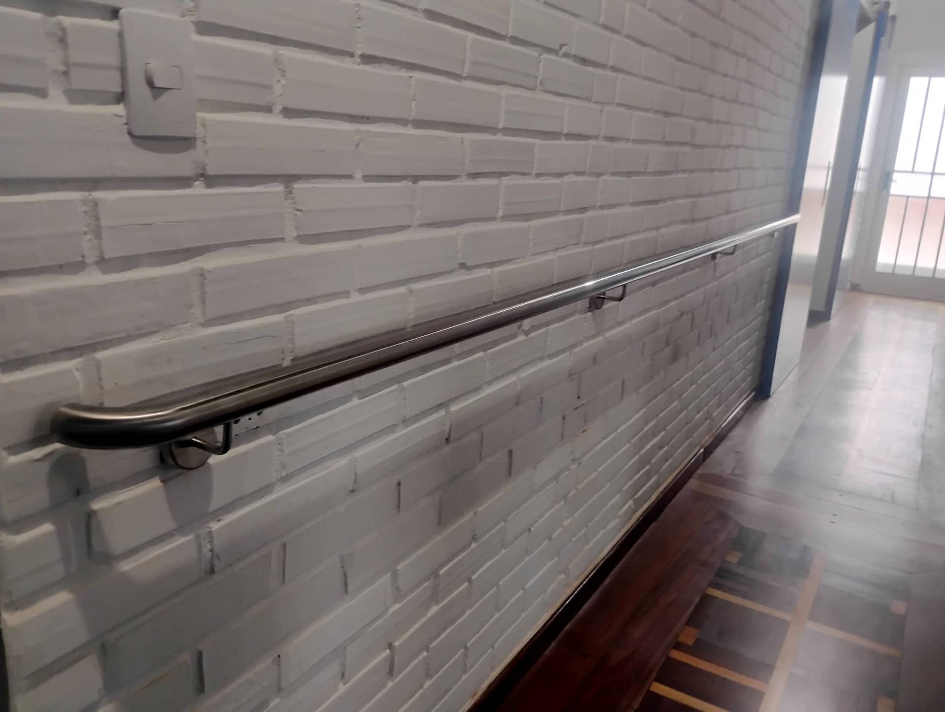 Corrimão em inox fixado em parede de tijolinho branco, ao longo do corredor