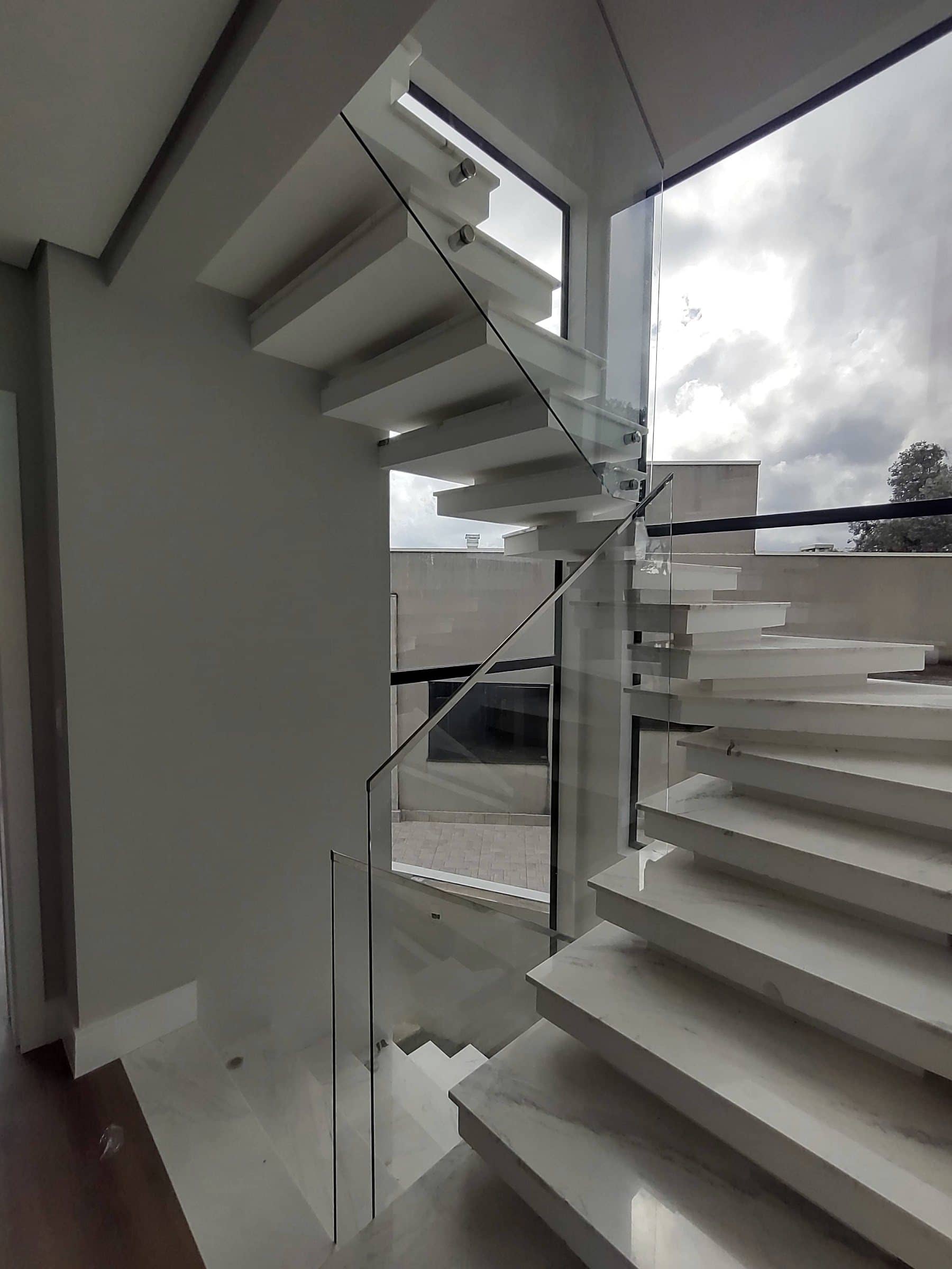 Escada em balanço vista de baixo, com vidro acompanhando os degraus junto à janela