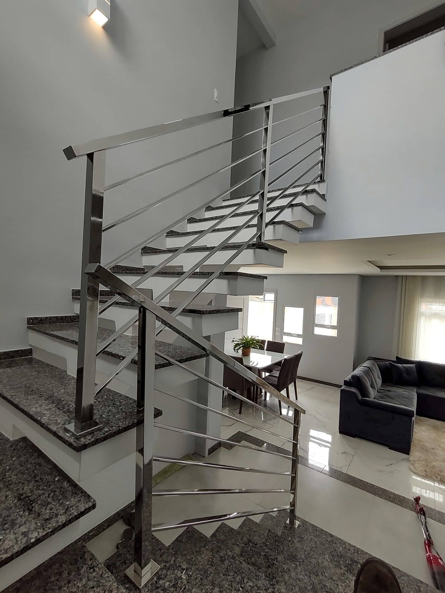 Escada com guarda-corpo de tubo quadrado em inox vista do pavimento superior, com a sala ao fundo