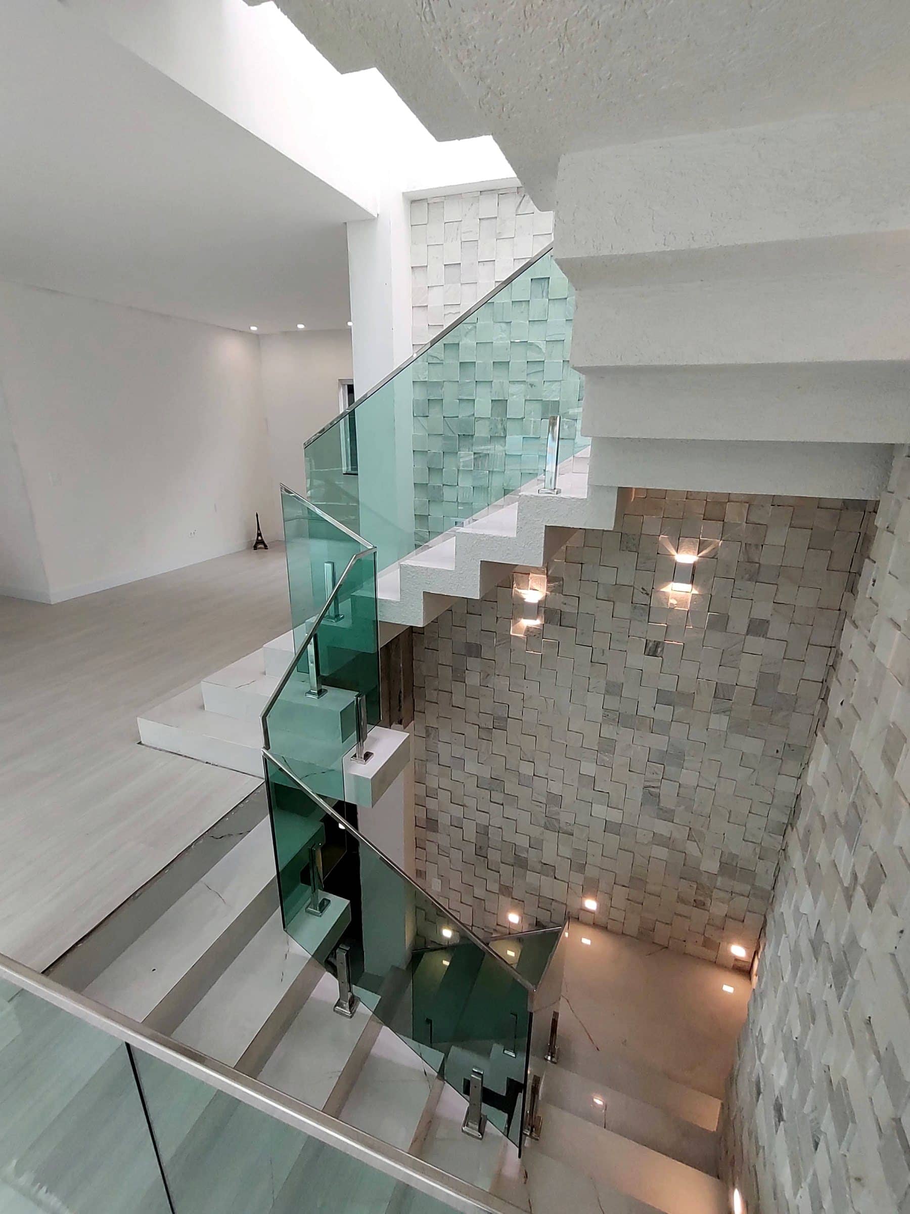 Escada de três pavimentos vista do alto, com guarda-corpo de vidro verde e colunas em inox ao lado de parede de pedra