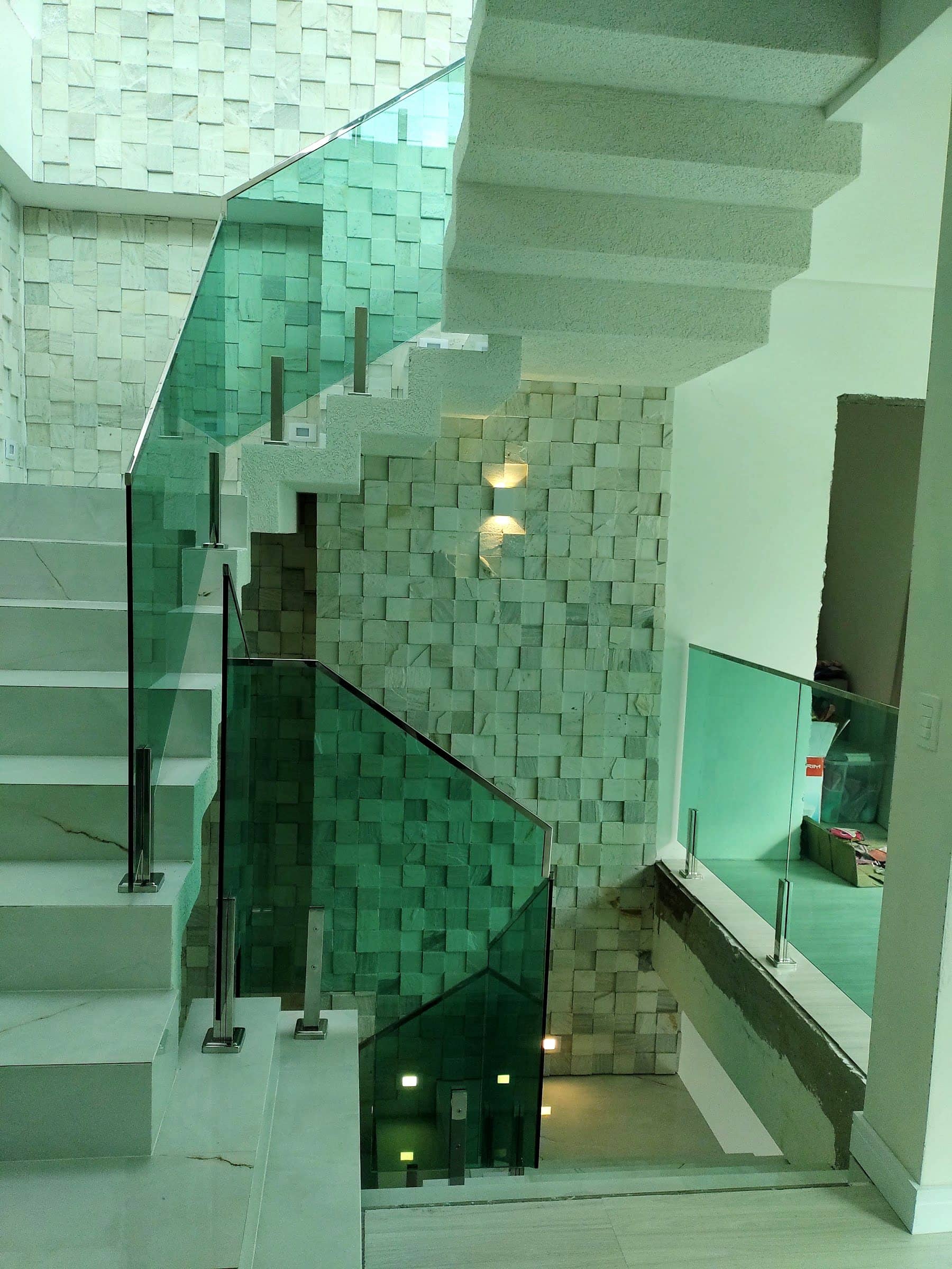 Guarda-corpo de vidro verde subindo junto à escada branca, com pinças em inox e parede de pedra iluminada