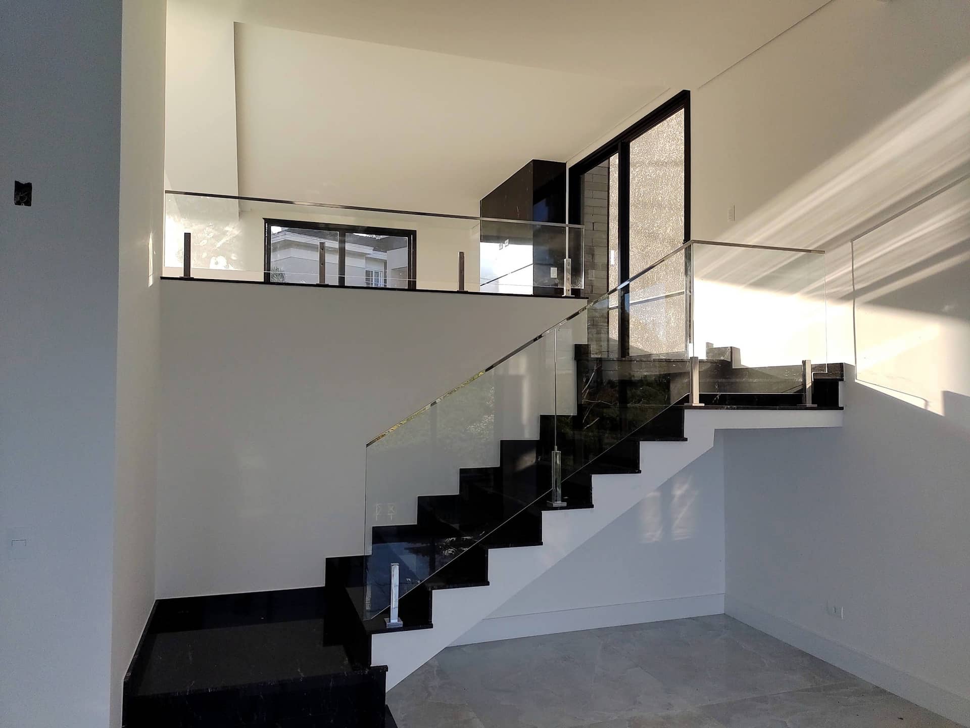 Escada em granito preto com guarda-corpo de vidro subindo até o mezanino