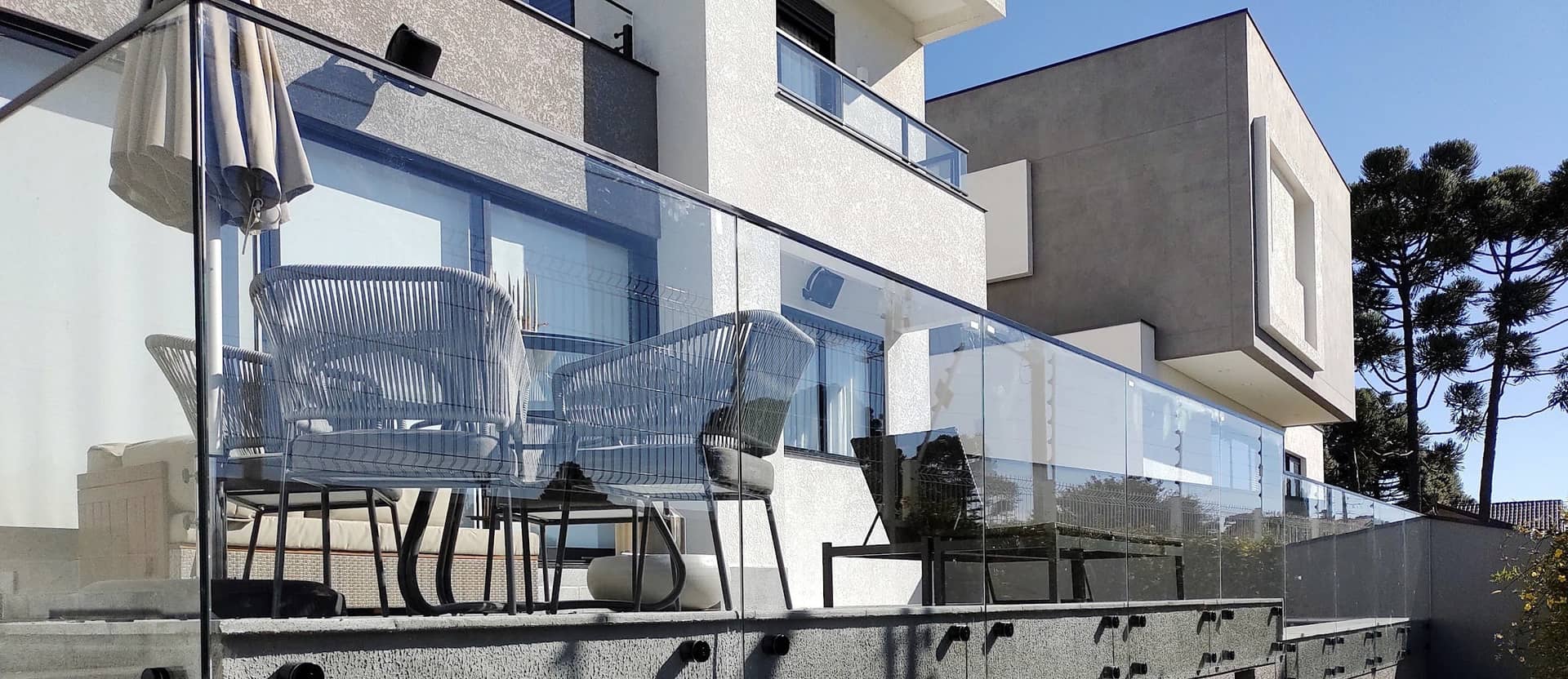 Terraço com guarda-corpo de vidro fixado por pinças em inox na lateral da laje, com araucárias ao fundo