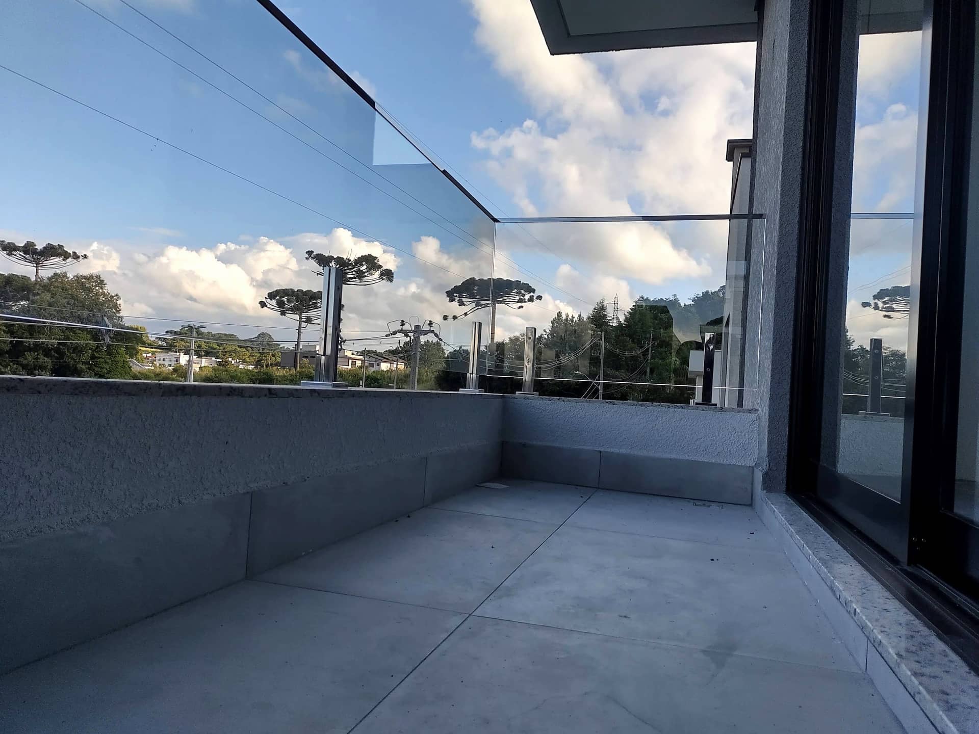 Varanda com guarda-corpo de vidro e vista para araucárias sob céu com nuvens